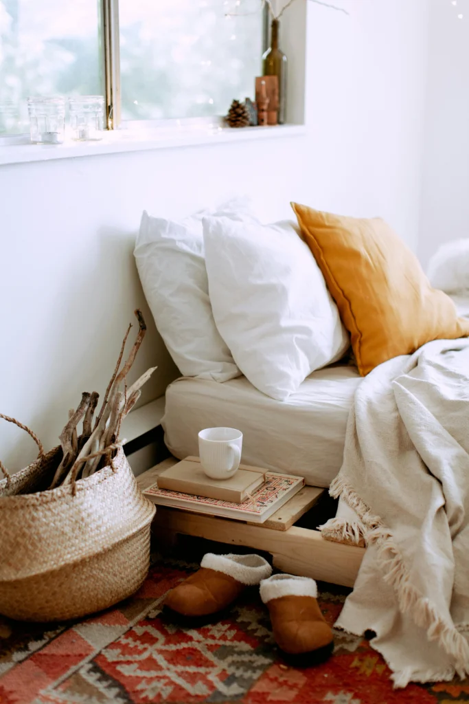 Image of a bed on a low wooden pallet frame, with white and mustard yellow pillows, a woven basket, and slippers.