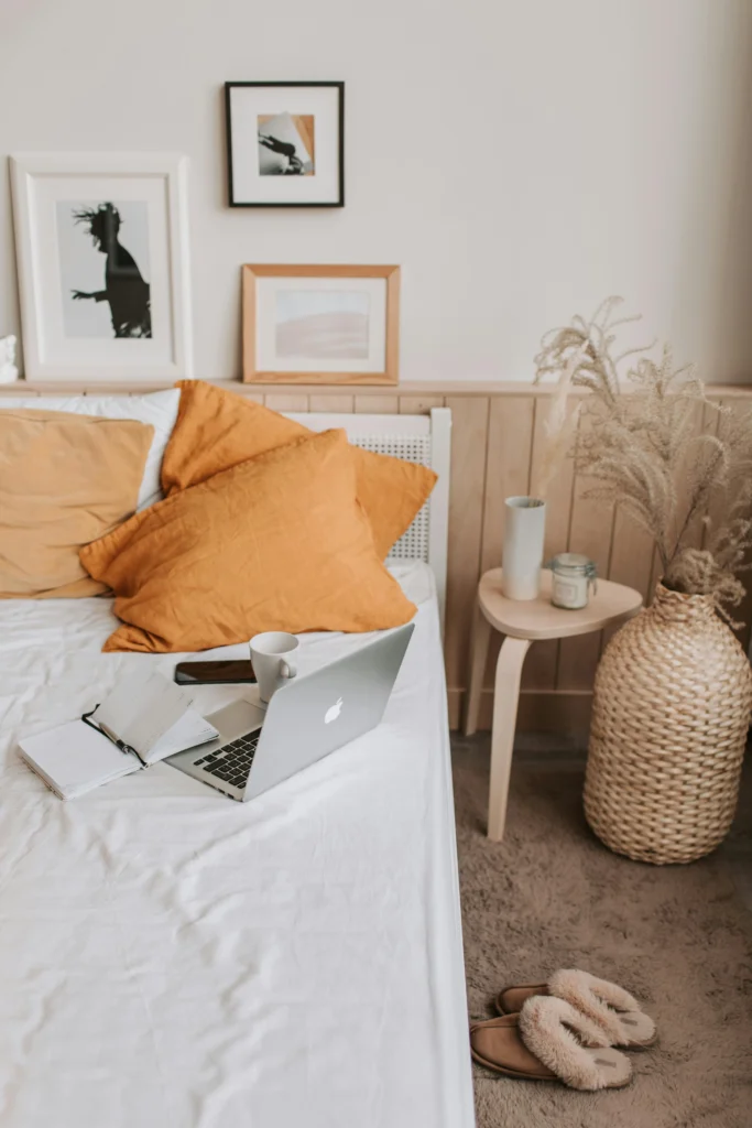 Image of a bed with white sheets and mustard yellow pillows, a laptop, and a bedside table with a large woven vase.