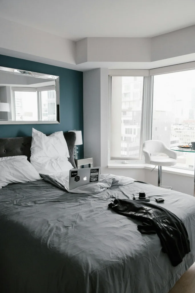 Image of bedroom with teal accent wall, large mirror, grey bedding, white chair.