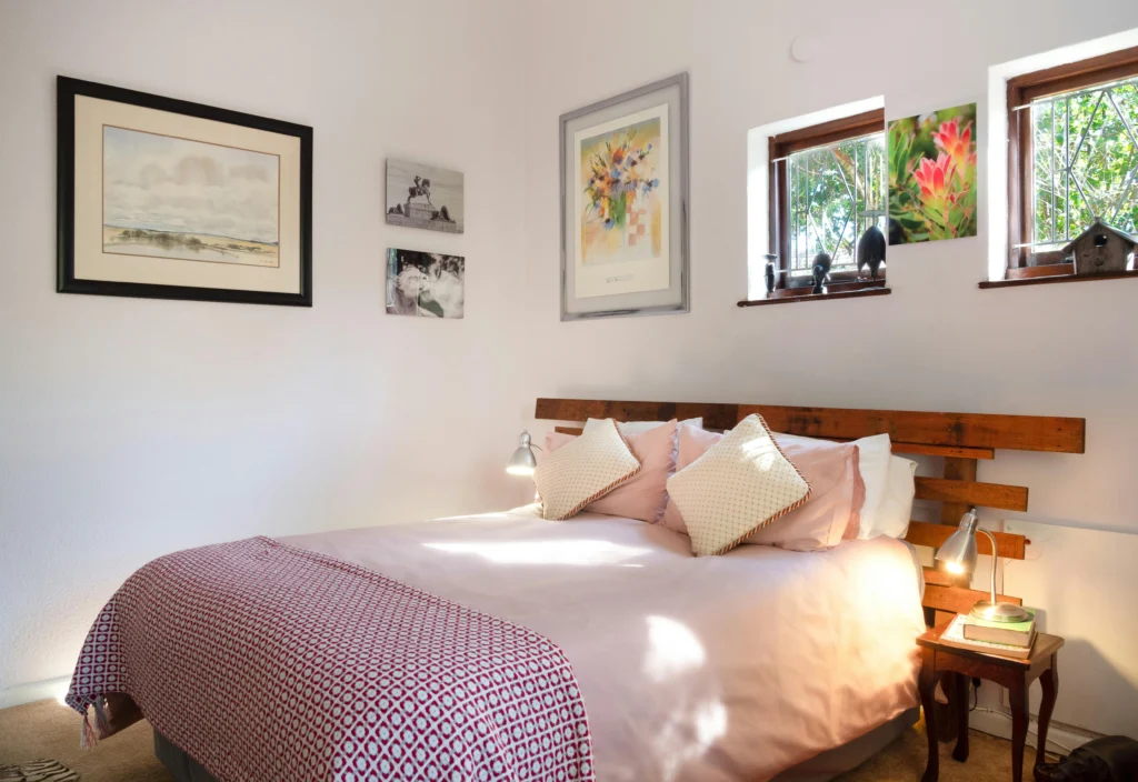 Image of bedroom with white walls, light wood headboard, pink bedding, framed artwork.