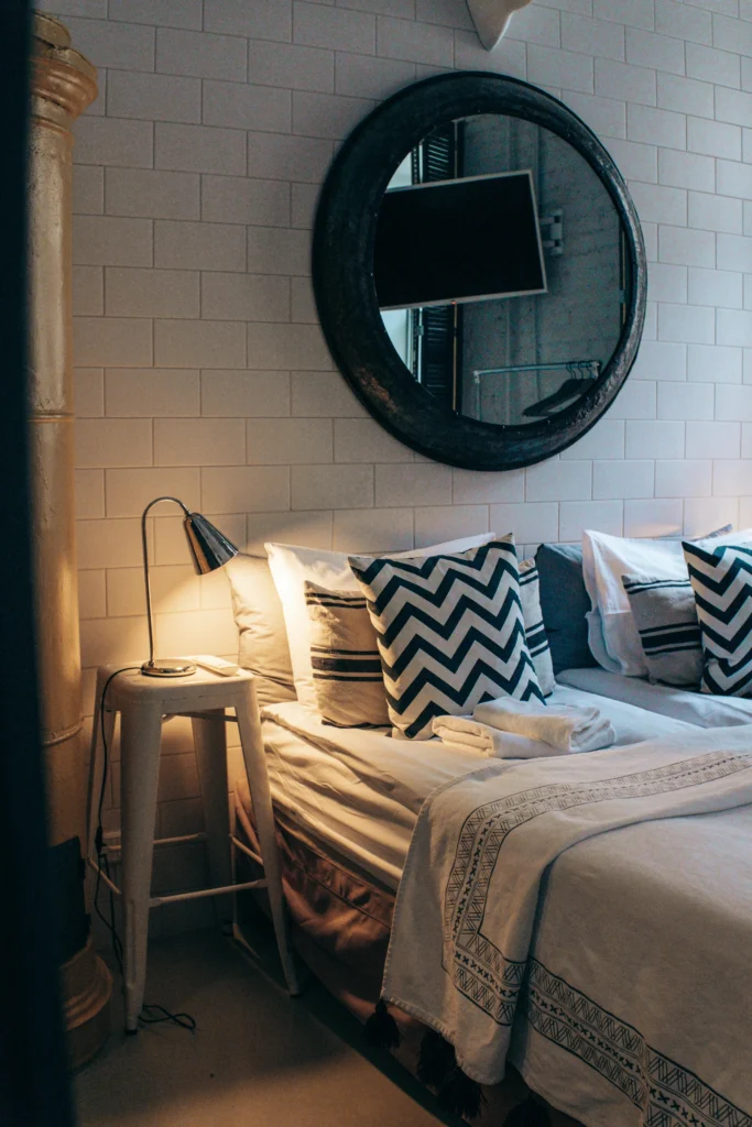 Image of bedroom with white brick wall, metal accents, patterned cushions, round mirror.